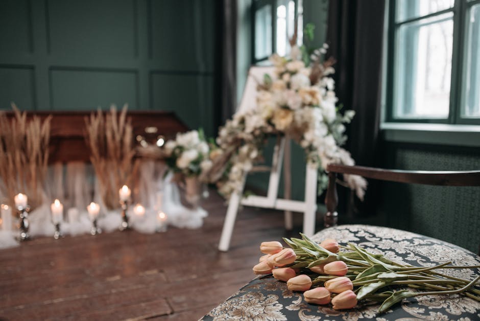 A floral arrangement featuring a bouquet of soft pink tulips with green stems and leaves resting on an ornate, patterned cushion. In the background, there is a white floral tribute adorned with white 