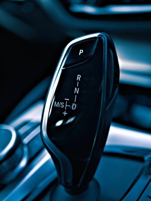 The image shows a close-up view of a modern automatic gear shift lever inside a vehicle. The gear lever features a sleek, black design with metallic accents, with visible markings indicating the vario