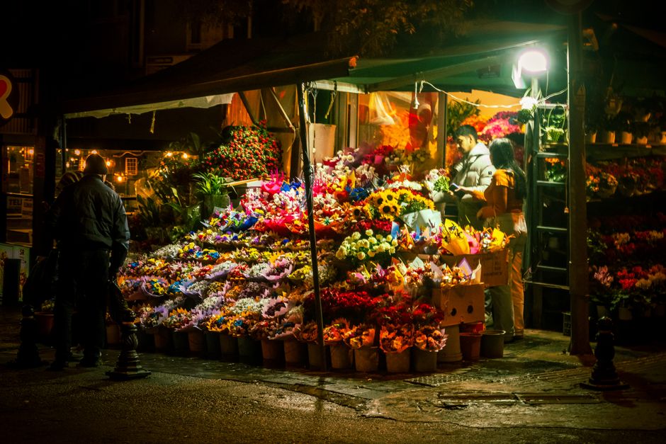 A nighttime street view of a flower stall on Barkingside High Street, illuminated by bright artificial lights. The stall is filled with a vibrant display of fresh flowers, including sunflowers, roses,