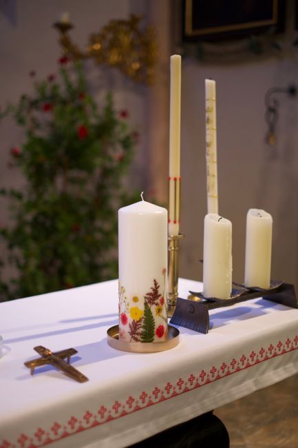 A collection of white candles, including a large pillar candle decorated with red, yellow, and green floral and foliage patterns, sits on a white cloth with a red cross-stitch border, arranged alongsi
