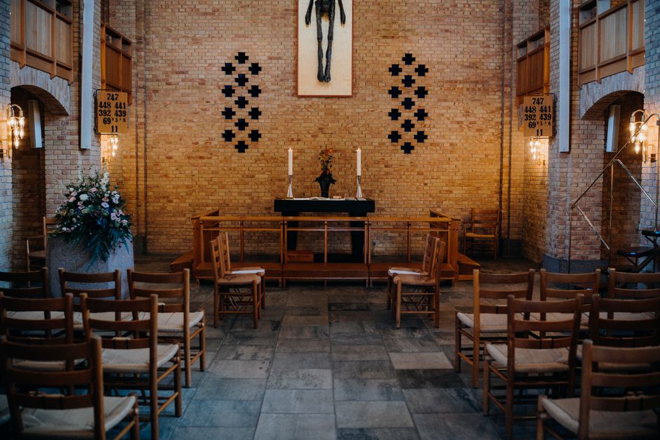 An interior view of a church decorated for a wedding, featuring a simple floral arrangement on a large urn with pink and white flowers and lush greenery, positioned on the left side near the brick arc