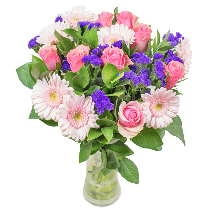 This vibrant yet gentle arrangement, expertly presented in a slender clear glass vase against a pure white backdrop, is a hallmark of Barkingside Florist's quality. The bouquet's focal point is a harmonious mix of 7-9 pink roses-some tightly budded, others unfolding into full, classic spirals-mirroring the romance and celebration often seen in Barkingside's local gatherings. Around them cluster 5-7 pale pink gerbera daisies, identifiable by their broad, plush heads and nearly-white central eyes that bring softness and balance. Deep purple statice, their stems arching through the bouquet, inject visual contrast and a lively pop of colour reminiscent of spring blooms at Barkingside's community gardens. Encasing these statement flowers, abundant, rich green foliage adds a sense of lushness and vibrancy, grounding the bouquet with the freshness of nearby clay soils. Visible stems and fresh water in the crystal-clear vase hint at careful local preparation and same-day Barkingside delivery. The bouquet's joyful blend of pinks and purples, balanced by green, makes it a fitting choice for birthdays, anniversaries, or simply brightening an office or front room in Barkingside, expressing heartfelt affection and the everyday artistry of a neighbourhood florist.